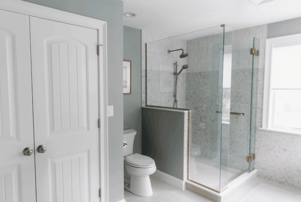 Corner view of glass-enclosed shower with champagne bronze hardware and toilet area in spa-inspired primary bathroom with soft blue-gray walls