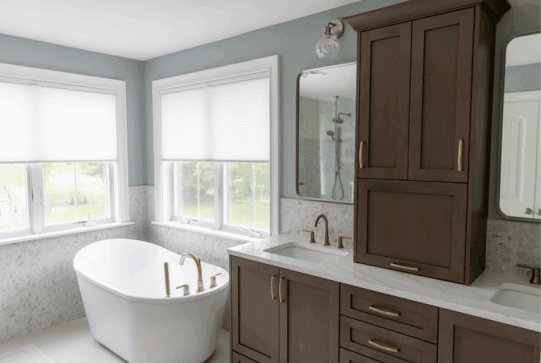 Freestanding white soaking tub with champagne bronze deck-mount faucet next to WoodHarbor cherry double vanity with rounded rectangle mirrors in serene primary bathroom
