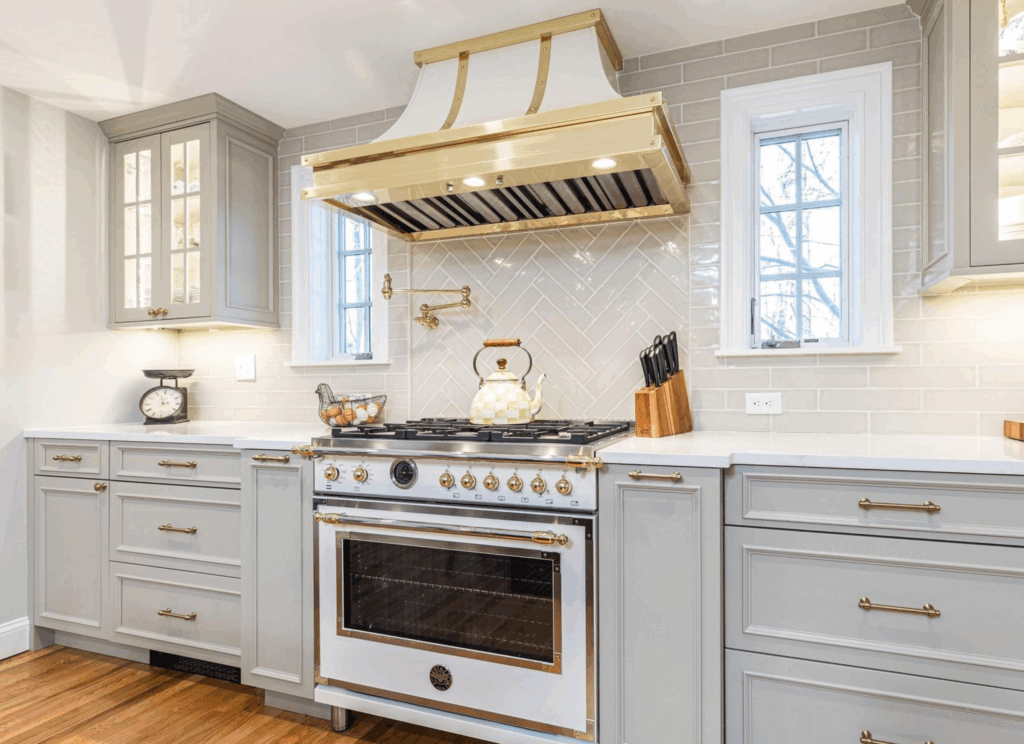 Custom gold Modern-Aire range hood and white Bertazzoni stove with gold accents, gray shaker cabinetry, white subway tile backsplash, and Calacatta Gold Silestone countertops in traditional kitchen design