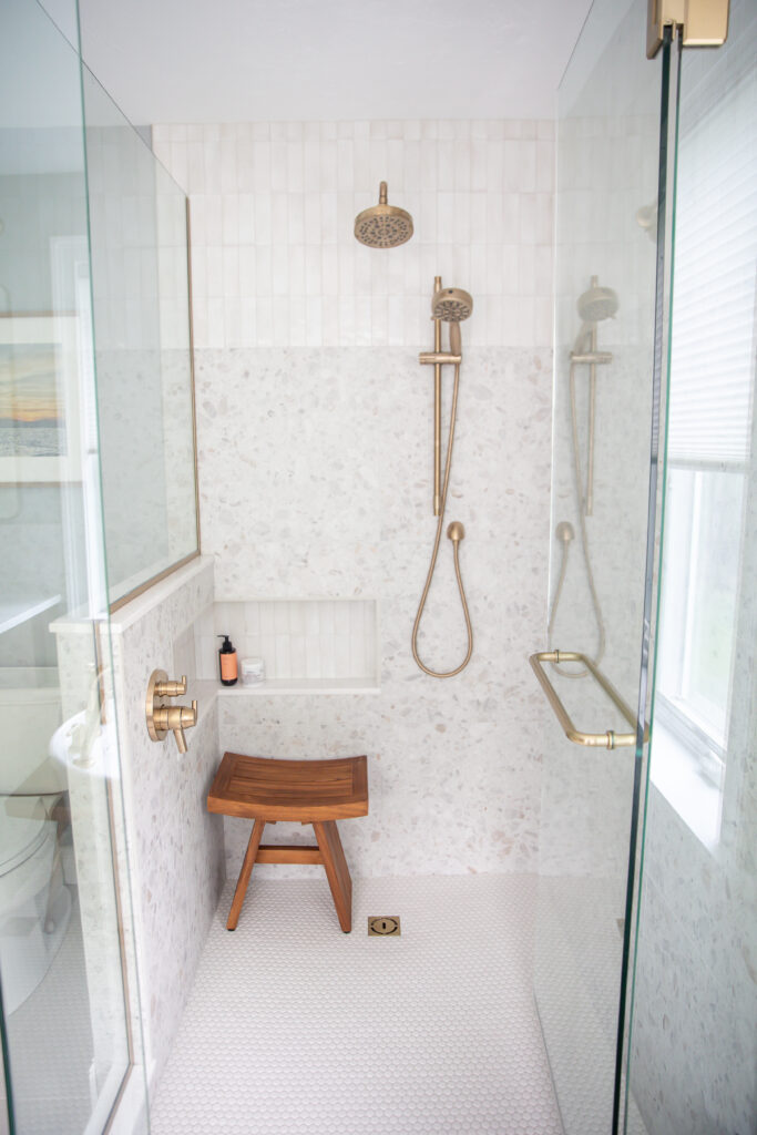 Glass shower enclosure with champagne bronze hardware, teak bench, terrazzo and white subway tile, and penny round flooring in resort-inspired primary bath