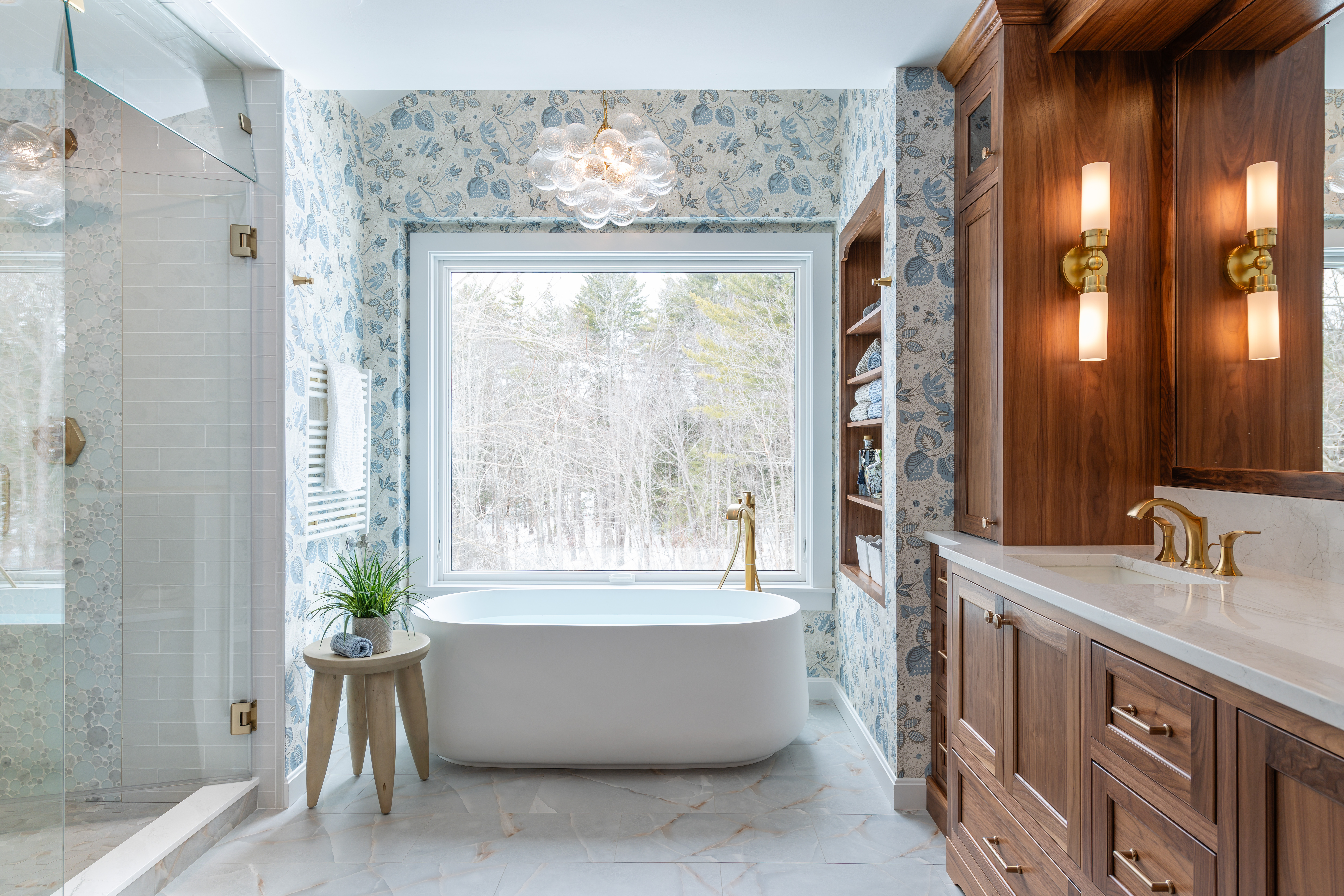 Primary bathroom featuring freestanding white tub beneath window with botanical blue wallpaper, custom walnut cabinetry with open shelving, marble tile steam shower with glass enclosure, and white countertops