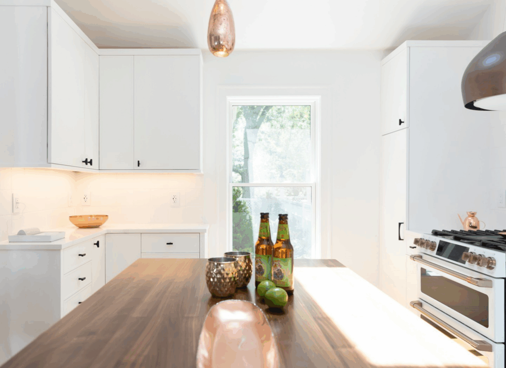 Custom Grothouse walnut wood countertop on white kitchen island with copper pendant lighting, white Café appliance with copper hardware, and natural light from Portsmouth home window