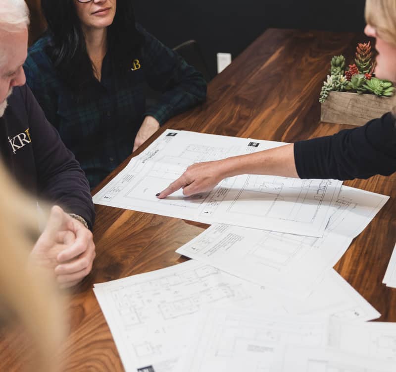 Close up of a hand pointing at design plans with two clients looking over the plans as well.